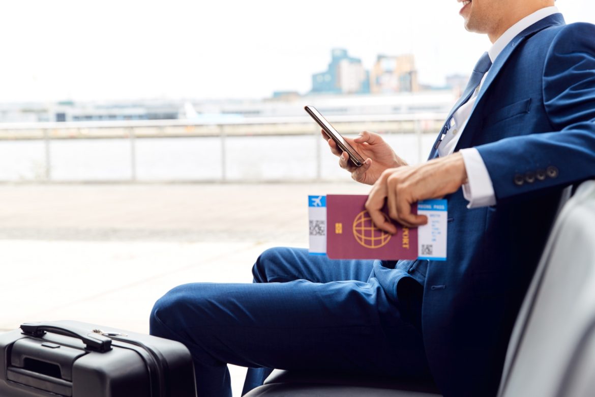 Businessman With Passport And Boarding Pass Sitting In Airport Departure Lounge Using Mobile Phone