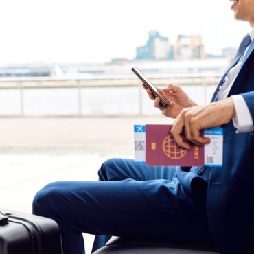 Businessman With Passport And Boarding Pass Sitting In Airport Departure Lounge Using Mobile Phone