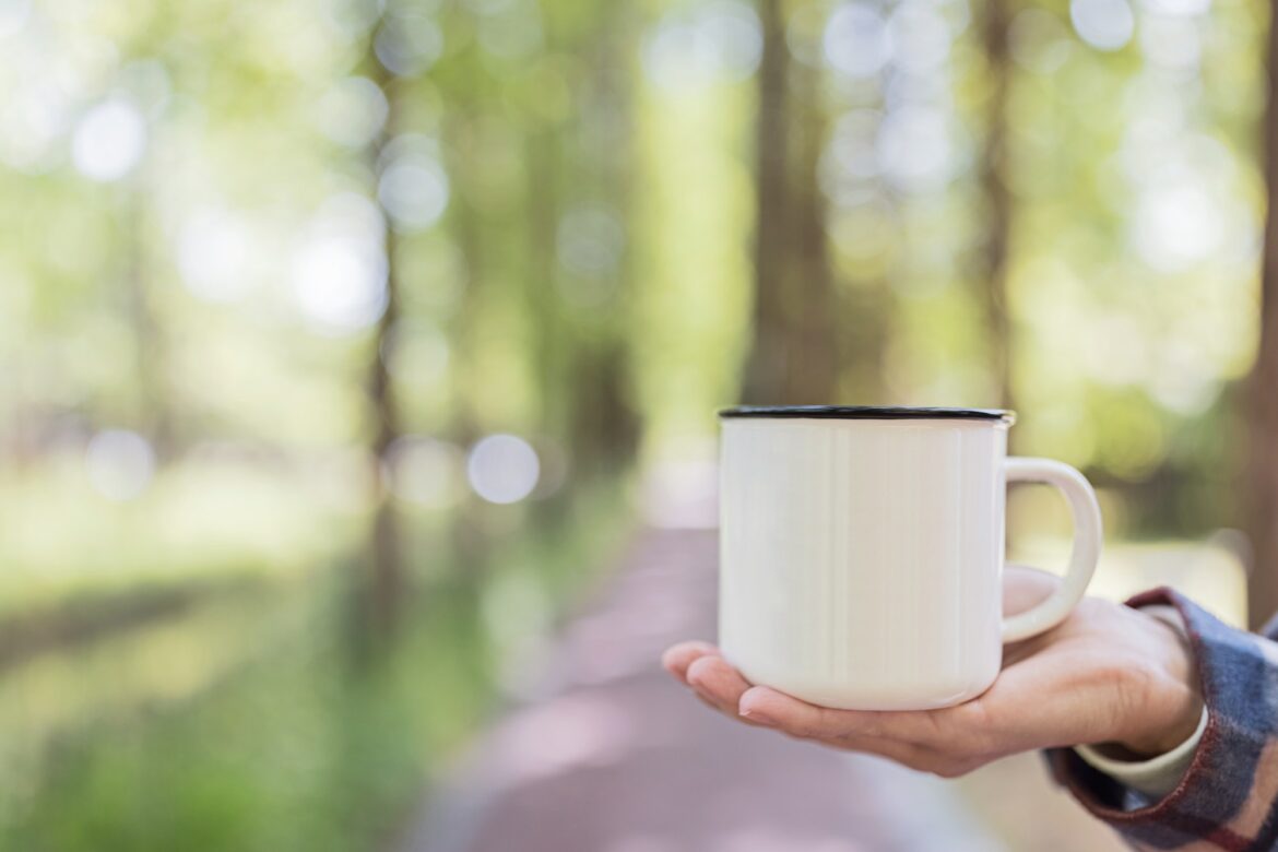 Woman hand holding metallic coffee mug in forest at early morning. Mockup with copy space