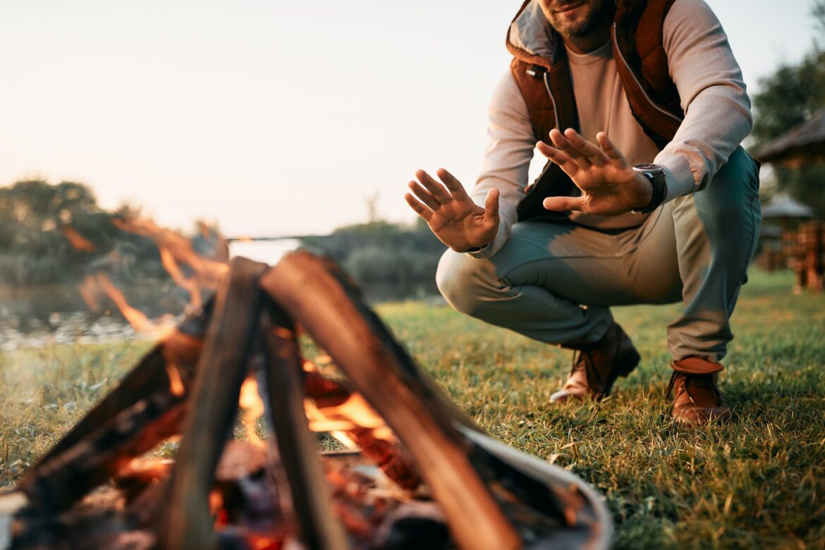 Unrecognizable man warming hands over campfire while camping in nature.