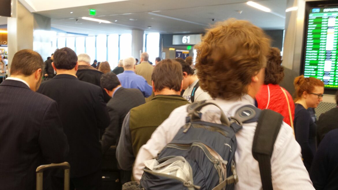 A crowded airport terminal with people standing in line. Some are wearing backpacks and carrying luggage. A digital departure board displaying flight information is visible on the right. The gate number 61 is seen in the background.