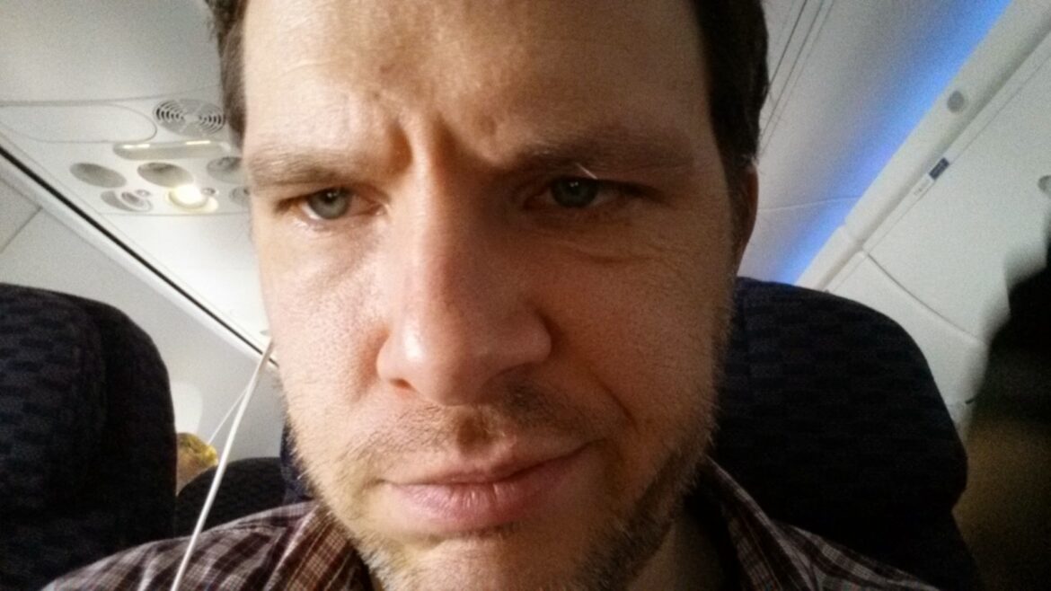 A close-up of a man sitting in an airplane. He has a serious expression and is wearing a plaid shirt. The airplane's interior, including seats and overhead compartments, is visible in the background.