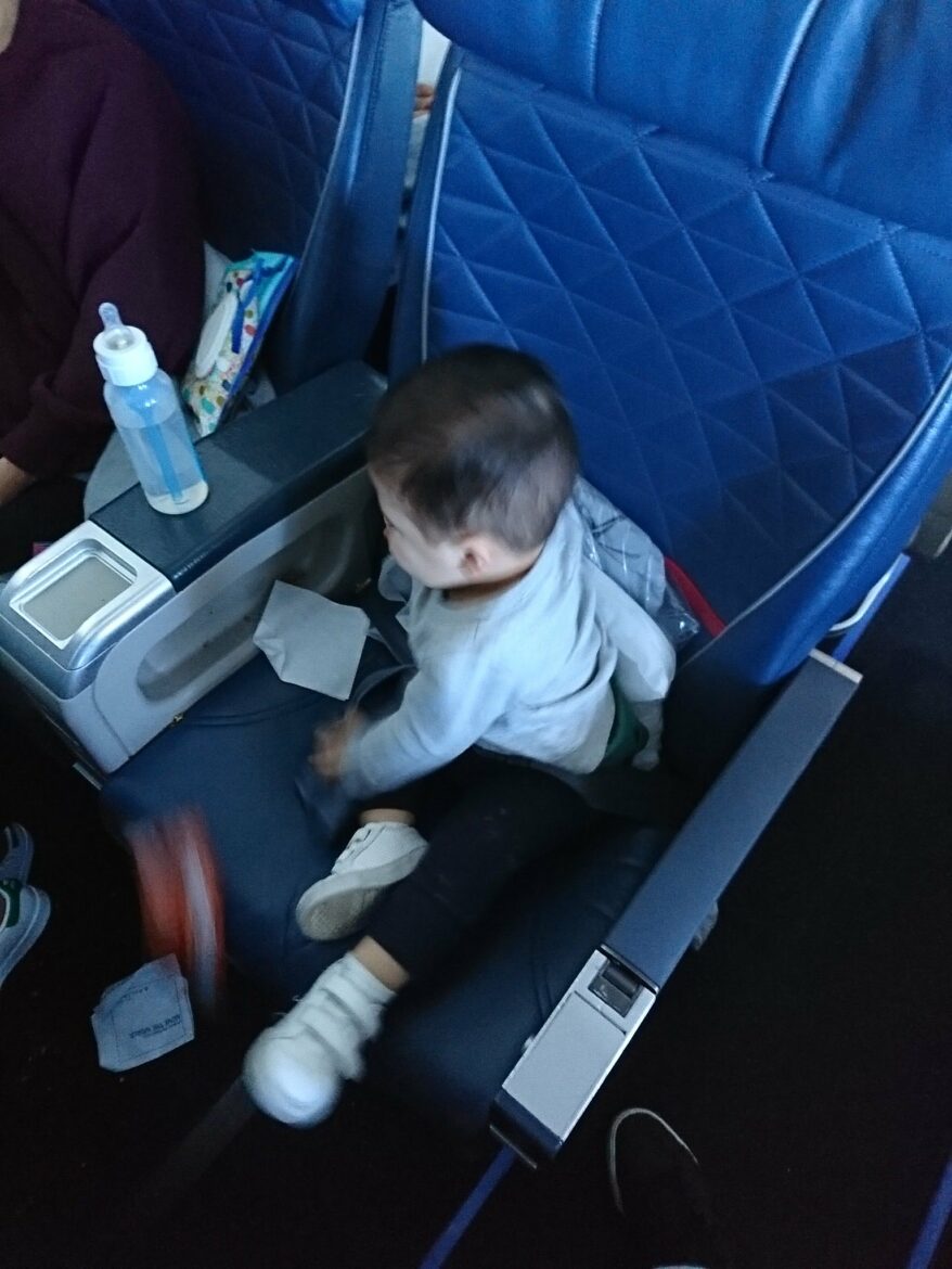 A small child is sitting on an airplane seat, wearing a light-colored shirt and dark pants. The child is playing with a toy and has one shoe on. The seat is blue with a quilted pattern. A bottle and some items are on the armrest. Another person is partially visible in the adjacent seat.