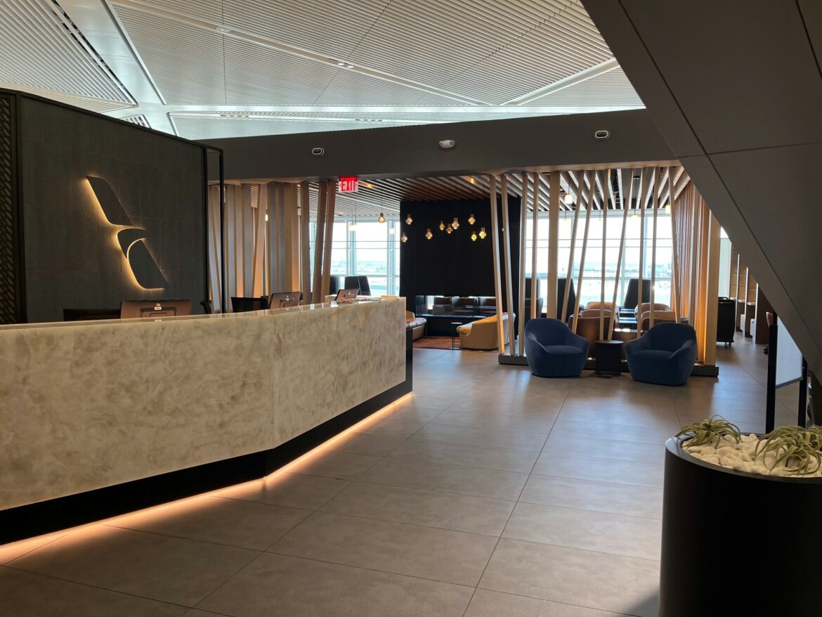 The image shows a modern airport lounge with a sleek, curved reception desk on the left. The desk is made of a light-colored material with subtle lighting underneath. Behind the desk, there are computer monitors and a wall with a stylized logo. To the right, there is a seating area with blue armchairs and a mix of other chairs and sofas. The space is well-lit with natural light coming through large windows, and there are decorative hanging lights. The ceiling has a geometric design, and there is a planter with white stones and plants in the foreground.