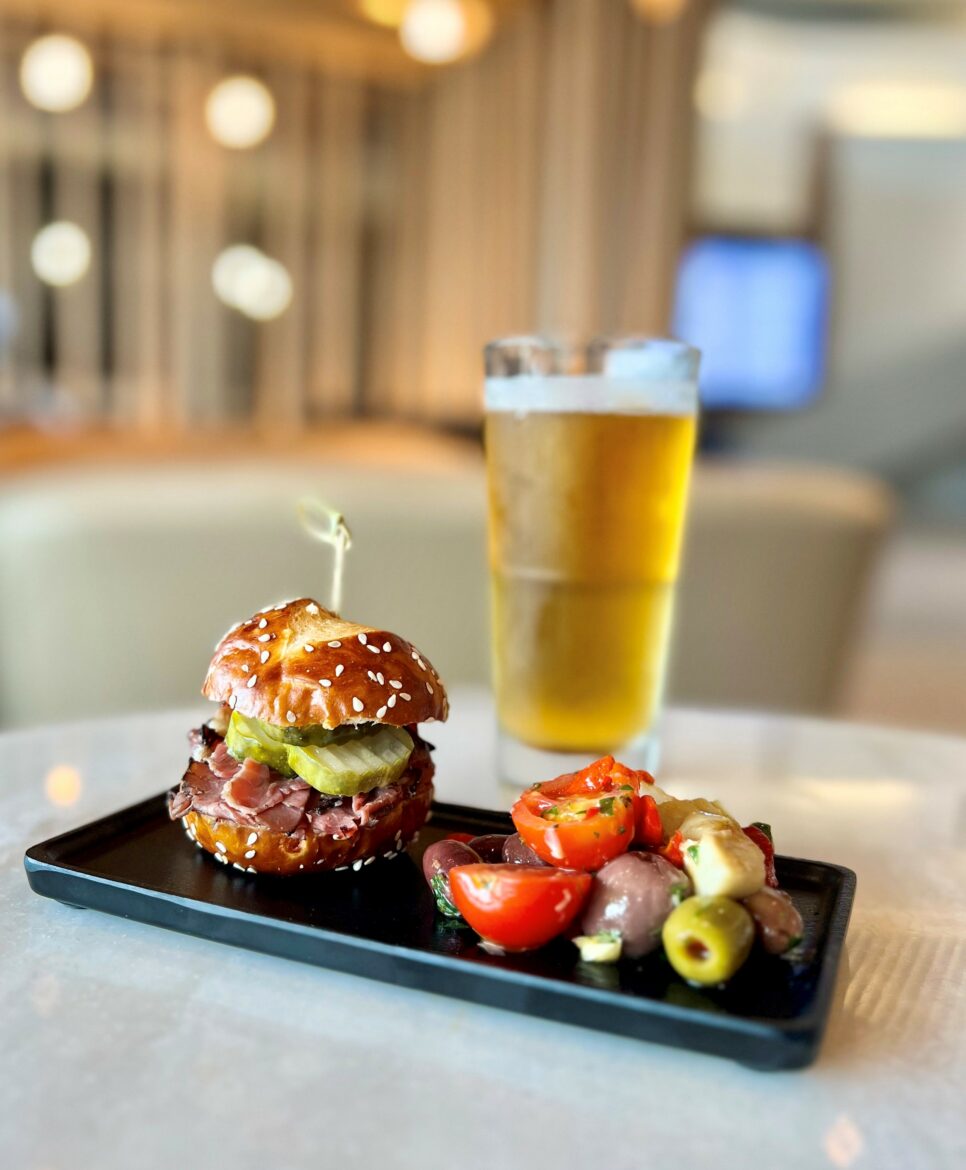 A small sandwich with sliced meat and pickles on a sesame seed bun is placed on a black rectangular plate. Next to it is a side of mixed olives and cherry tomatoes. In the background, there is a tall glass of beer. The setting appears to be a casual dining area with soft lighting.