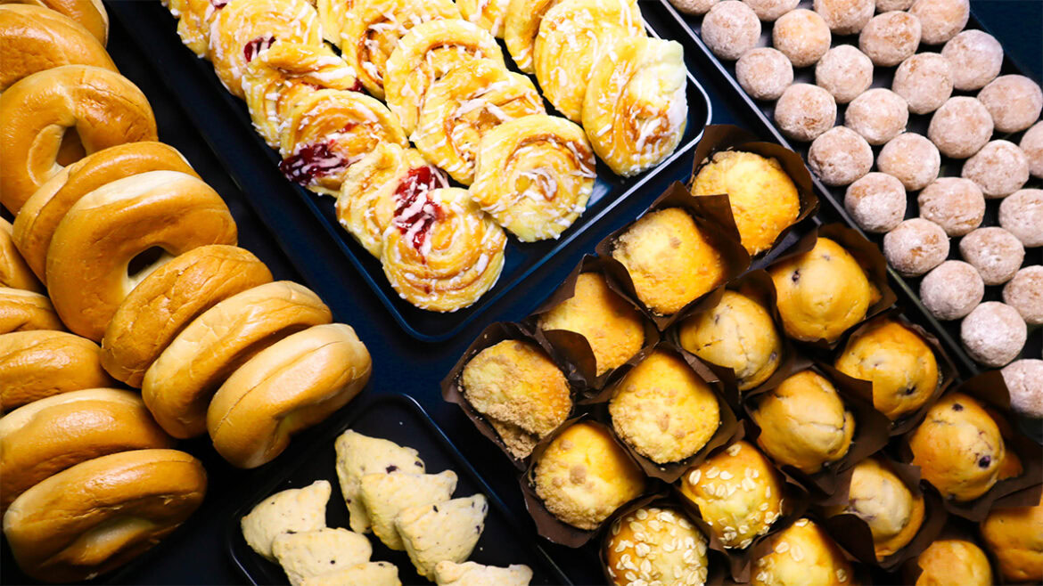 The image shows an assortment of baked goods arranged on trays. There are bagels on the left, pastries with icing and fruit filling in the center, muffins in brown paper cups, and rows of small round doughnut holes or cookies on the right. The items are displayed on a dark surface.