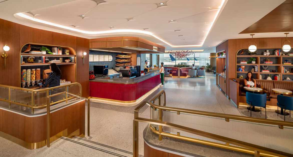 The image shows a modern airport lounge with a stylish interior. In the foreground, a woman is using a snack dispenser area with various snacks like candy and nuts. To the right, another woman is seated at a booth with a drink. The lounge features wooden paneling, bookshelves with decorative items, and a red counter where a person is being served by a staff member. In the background, there are more seating areas with people relaxing, and large windows offering a view of the airport tarmac. The space is well-lit with contemporary lighting fixtures.