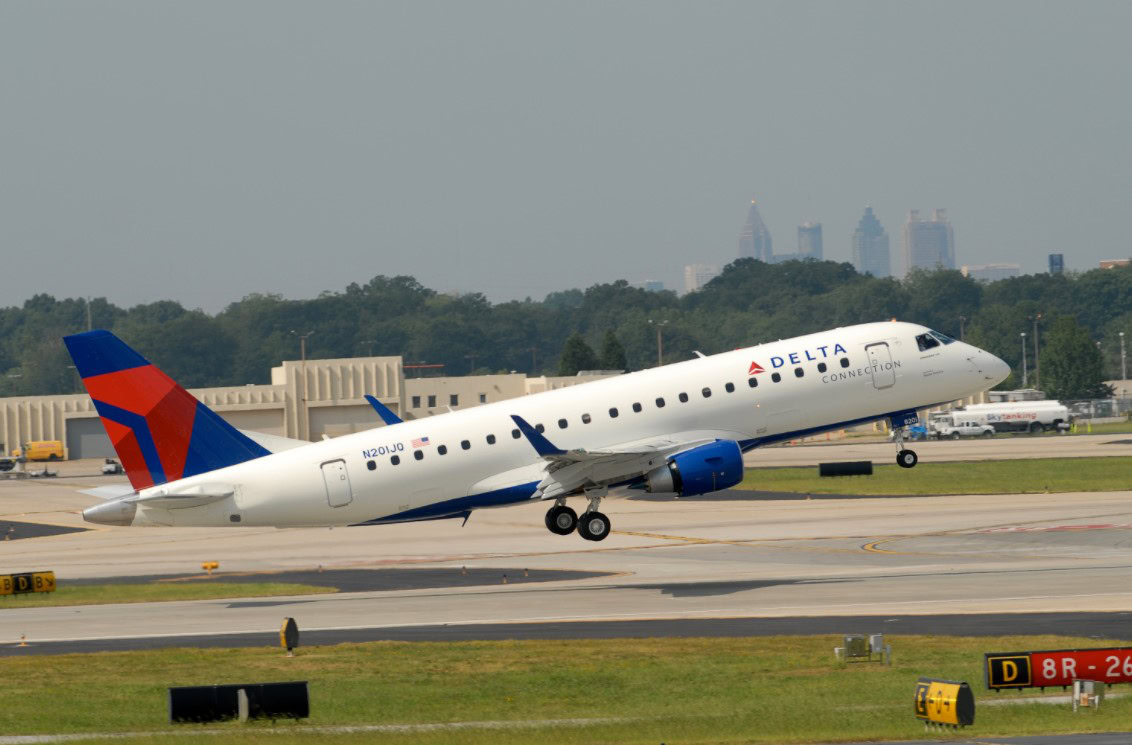 A Delta Connection airplane is taking off from a runway at an airport. The aircraft is angled upwards, with its landing gear still extended. The background shows airport buildings, trees, and a city skyline under a clear sky.