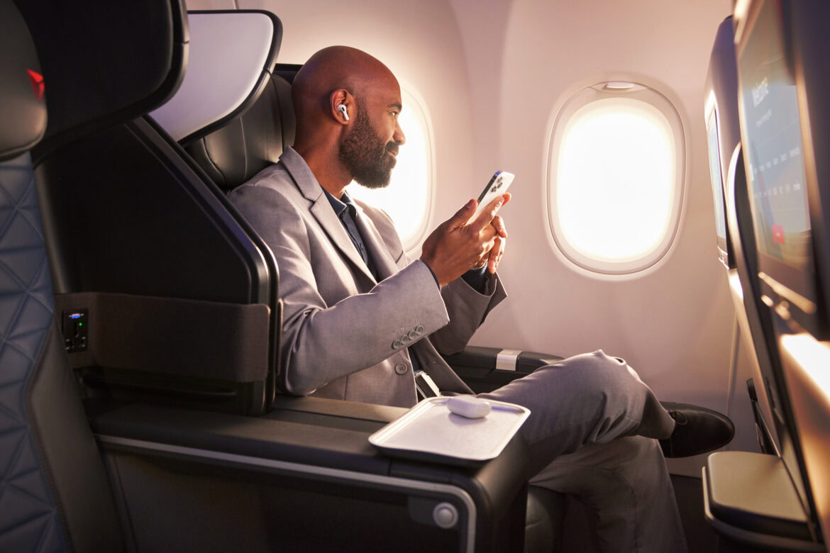 A man in a suit is sitting in a first-class airplane seat, holding a smartphone. He is wearing wireless earbuds and appears relaxed. The seat has a tray with a small item on it, and there is a window with sunlight streaming in. A screen is visible in front of him.