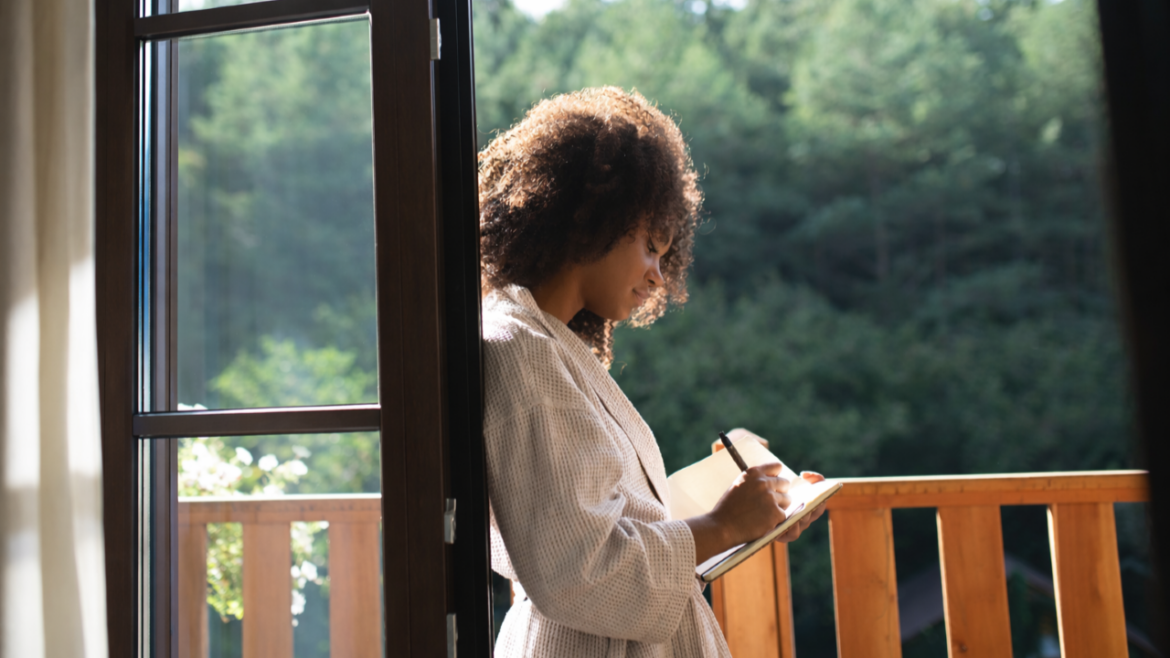 A person with curly hair is standing on a balcony, writing in a notebook. They are wearing a light-colored robe and are partially illuminated by sunlight. The background shows a view of green trees.