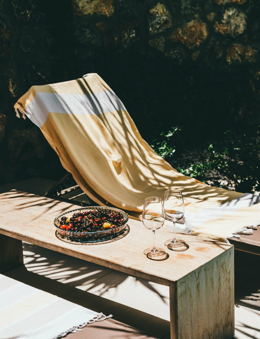 A sunlit outdoor setting featuring a wooden lounge chair covered with a yellow and white towel. In front of the chair is a wooden table with a bowl of cherries and two empty wine glasses. Shadows of leaves are cast on the scene, and a stone wall is visible in the background.