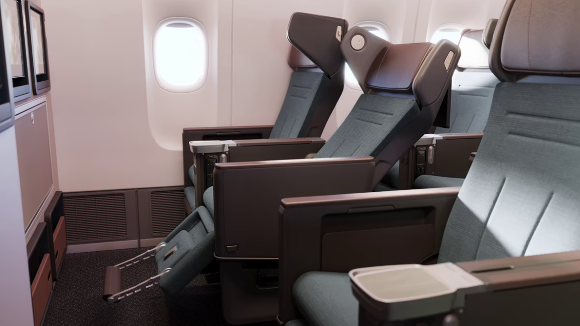 The image shows a row of premium economy or business class airplane seats. The seats are spacious with adjustable headrests and footrests. Each seat has a personal entertainment screen and a tray table. The cabin interior is modern and well-lit, with a window visible in the background.