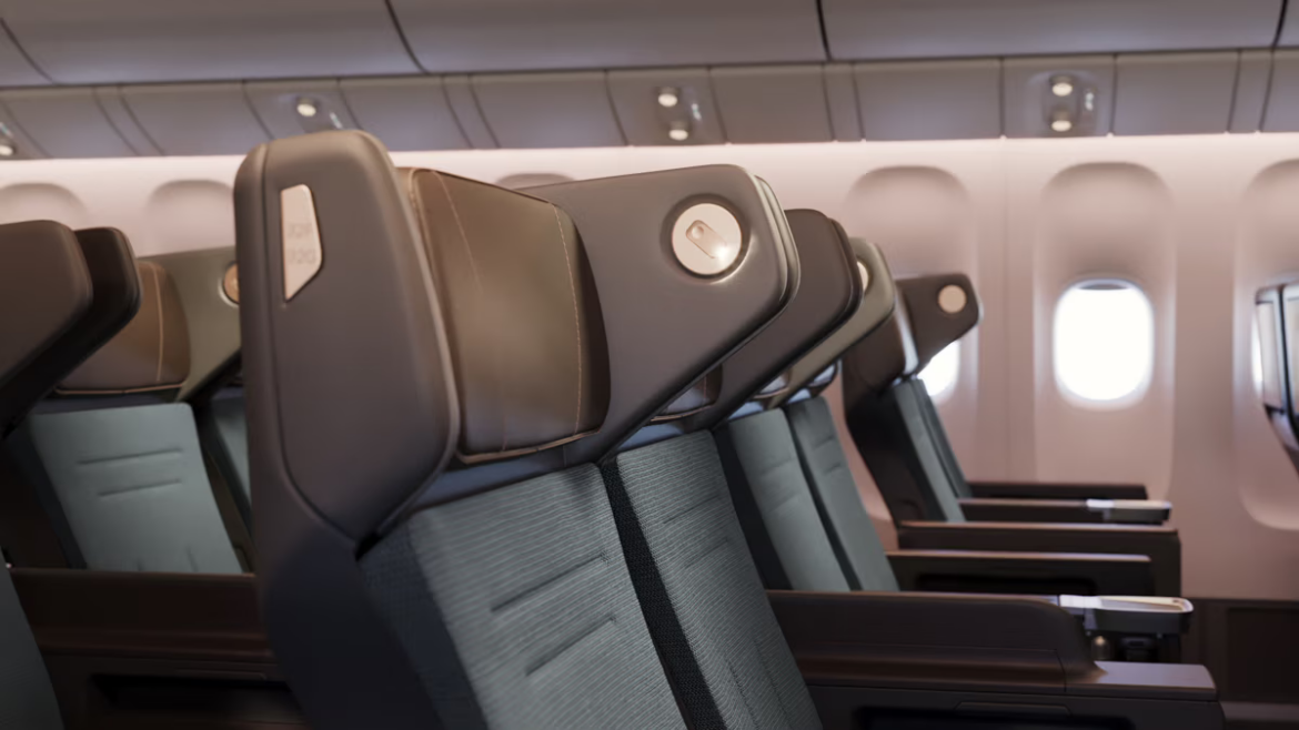 The image shows a row of modern airplane seats in an aircraft cabin. The seats have dark upholstery with sleek headrests and are arranged in a typical economy class configuration. The cabin interior is well-lit, and airplane windows are visible in the background.