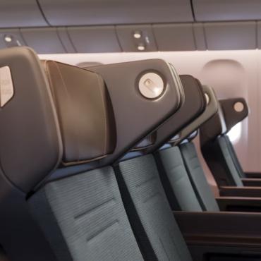 The image shows a row of modern airplane seats in an aircraft cabin. The seats have dark upholstery with sleek headrests and are arranged in a typical economy class configuration. The cabin interior is well-lit, and airplane windows are visible in the background.
