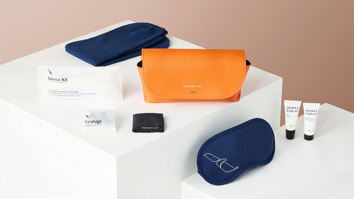 The image shows a collection of travel amenities arranged on white blocks against a beige background. Items include a blue eye mask, a pair of blue socks, an orange pouch labeled "Raven + Lily," a dental kit, earplugs, a small black cloth, and two tubes of Joanna Vargas skincare products.