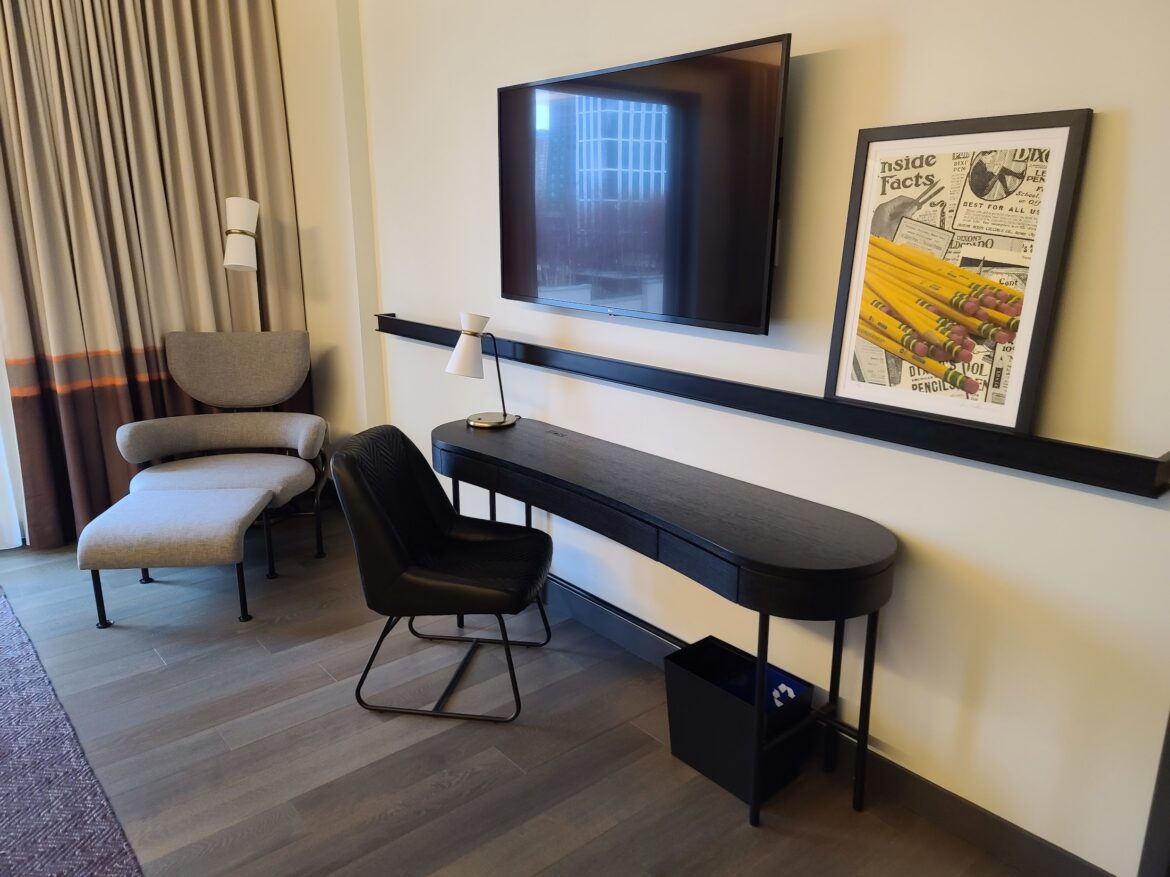 The image shows a modern hotel room corner with a flat-screen TV mounted on the wall. Below the TV is a sleek black desk with a small white lamp and a black chair. To the right of the desk, there is a framed artwork featuring yellow pencils and newspaper clippings. On the left side, there is a gray upholstered chair with a matching ottoman, accompanied by a floor lamp. The room has wooden flooring and curtains in shades of beige and brown.