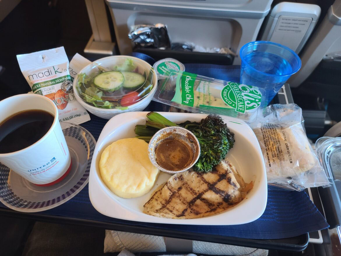 The image shows an airplane meal on a tray table after upgrading to premium economy. The meal includes grilled chicken, broccoli, a small round portion of mashed potatoes, and a sauce in a small container. There is also a side salad with cucumber slices, a packet of almonds, a chocolate chip cheesecake, and a wrapped bread roll. A cup of black coffee and a blue plastic cup of water are also present. The setting is inside an airplane cabin.