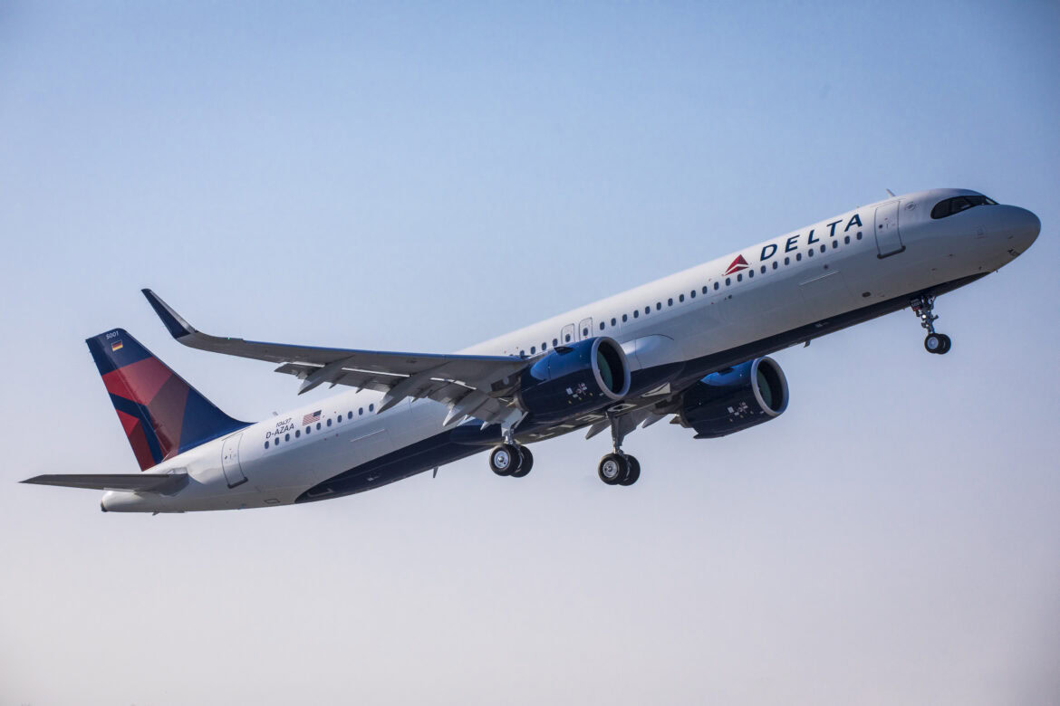 The image shows a Delta Air Lines airplane in flight against a clear blue sky. The aircraft is angled upwards, suggesting it is taking off or climbing. The Delta logo is visible on the side of the plane and on the tail fin.