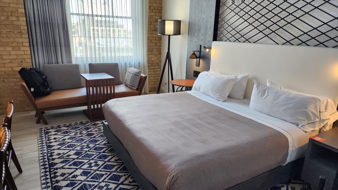 A modern hilton diamond reserve upgrade with a large bed featuring white pillows and a gray bedspread. To the left, there is a seating area with a brown cushioned bench, gray pillows, and a small wooden table. A black backpack is on the bench. A floor lamp stands next to the bed, and a round wooden side table is nearby. The wall behind the bed has a geometric black lattice design. A large window with sheer curtains allows natural light into the room, and a patterned blue and white rug is on the floor.