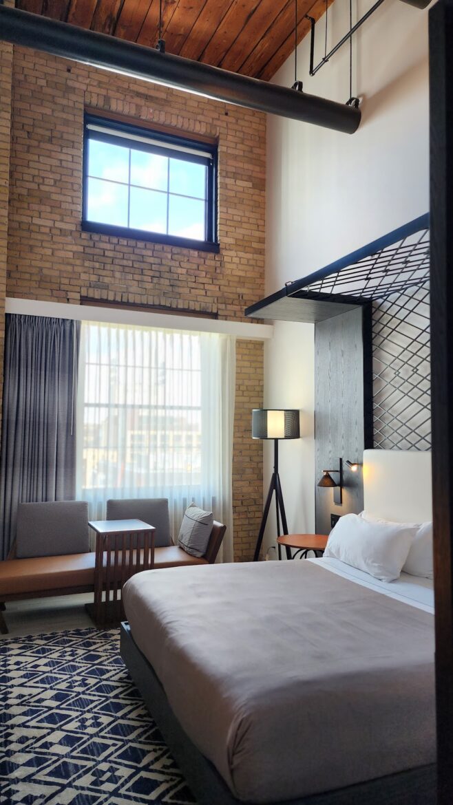 A hilton diamond reserve upgrade room with a high ceiling and exposed brick walls. The room features a large bed with white linens, a patterned rug, and a seating area with a small table and chairs. There is a tall floor lamp next to the bed and a large window with sheer curtains, allowing natural light to fill the space. The ceiling has wooden beams and a visible duct.