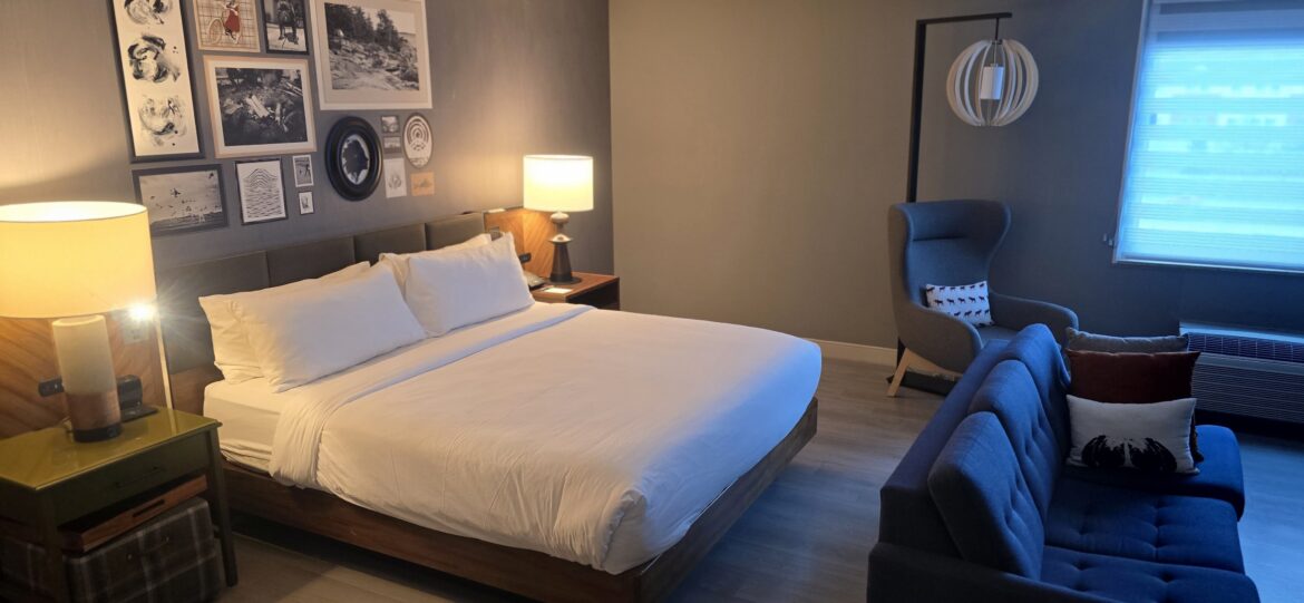 A Marriott free night certificate top off could find you a cozy hotel room with a large bed featuring white linens and pillows. The room has a modern design with a wooden headboard and two bedside lamps. The wall above the bed is decorated with various framed artworks. There is a blue sofa with decorative pillows and a gray armchair with a patterned pillow. A window with blinds allows natural light to enter the room.