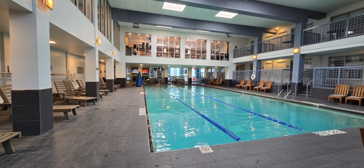 Swim up with your Marriott free night certificate top off. The image shows an indoor swimming pool with clear blue water, divided into lanes by floating lane markers. Surrounding the pool are lounge chairs and a tiled floor. The area is well-lit with ceiling lights, and there are two levels of seating or viewing areas with glass windows. The space has a modern design with white walls and dark accents.