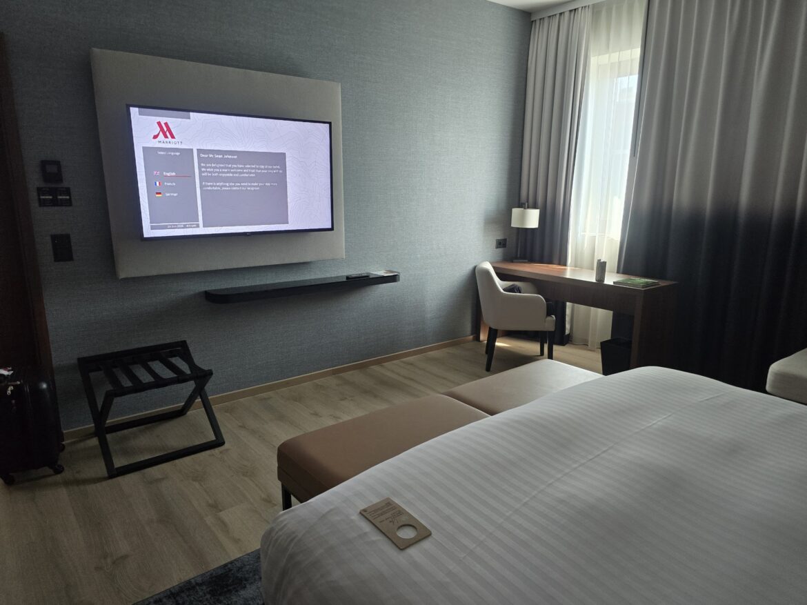 The image shows a modern hotel room with a large bed in the foreground. Teh rusult of a Marriott free night certificate top off. On the wall, there is a flat-screen TV displaying a welcome message. Below the TV is a small shelf. To the right, there is a wooden desk with a white chair, a lamp, and a window with sheer and blackout curtains. The floor is wooden, and there is a luggage rack near the TV.