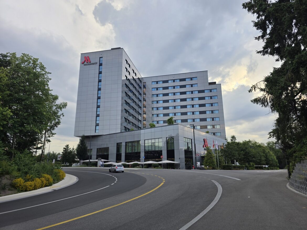 The image shows a modern multi-story hotel building with a sleek glass and metal exterior. The hotel has a prominent logo on the top corner. In front of the building, there is a curved driveway with a car parked near the entrance. Several flags are displayed near the entrance, and there are trees and greenery surrounding the area. The sky is cloudy, suggesting an overcast day. The benefits of your Marriott free night certificate top off.