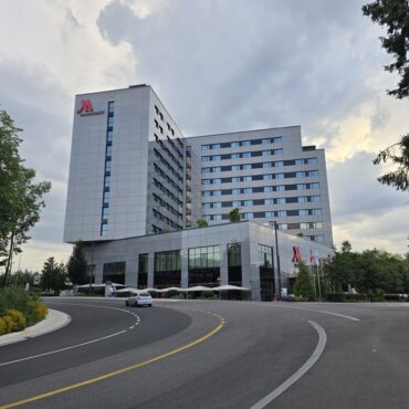 The image shows a modern multi-story hotel building with a sleek glass and metal exterior. The hotel has a prominent logo on the top corner. In front of the building, there is a curved driveway with a car parked near the entrance. Several flags are displayed near the entrance, and there are trees and greenery surrounding the area. The sky is cloudy, suggesting an overcast day. The benefits of your Marriott free night certificate top off.