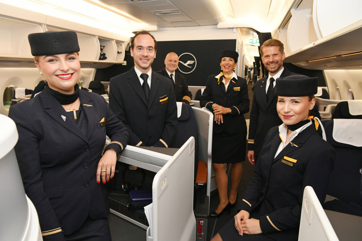 A group of six airline crew members, dressed in formal uniforms, are smiling and posing inside an airplane cabin. The cabin appears modern and well-lit, with visible seating and overhead compartments. The crew members are wearing matching dark uniforms with gold accents and hats.