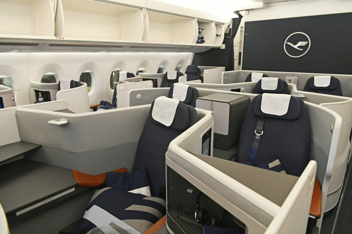 The image shows the interior of an airplane's business class cabin. The seats are arranged in a staggered configuration, providing privacy and space for each passenger. Each seat has a personal entertainment screen, a pillow, and a blanket. The cabin is well-lit, with overhead compartments for storage. The airline's logo is visible on the wall at the front of the cabin.