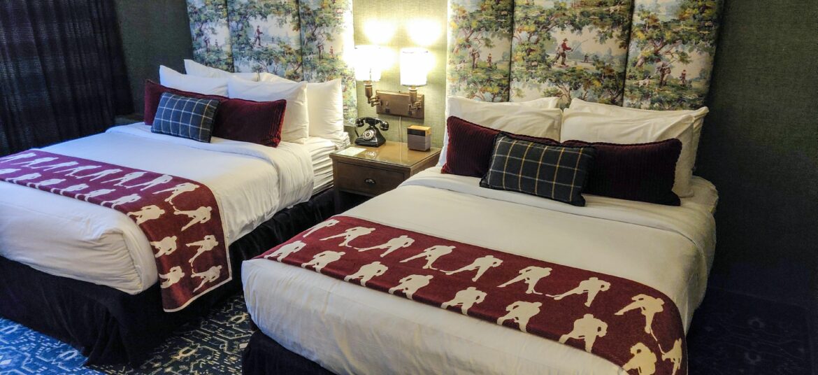 The image shows a hilton diamond reserve upgrade with two neatly made double beds. Each bed has white linens, burgundy throw pillows, and a decorative runner featuring silhouettes of hockey players. A nightstand with a vintage-style telephone and a lamp is positioned between the beds. The headboard is upholstered with a scenic fabric depicting people in a park setting. The room has a cozy and classic decor.