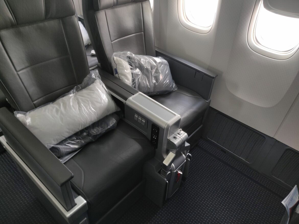 The image shows two adjacent airplane seats after upgrading to premium economy. The seats are upholstered in dark leather and have armrests with control panels. Each seat has a packaged pillow and blanket placed on it. There are two windows visible on the aircraft wall next to the seats.