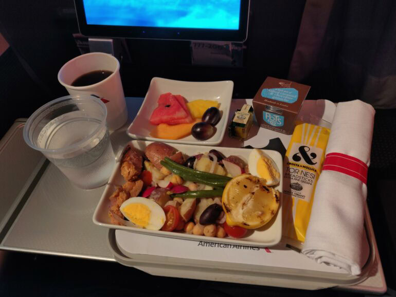 The image shows an airline meal on a tray table. The meal includes a salad with ingredients like boiled eggs, green beans, potatoes, chickpeas, and grilled lemon. There is a side dish of sliced watermelon, cantaloupe, and grapes. Accompanying the meal are a cup of coffee, a plastic cup of water, a small container of butter, a dessert labeled "Pots & Co," and a package of Torinesi breadsticks. A rolled-up napkin is also present on the tray.