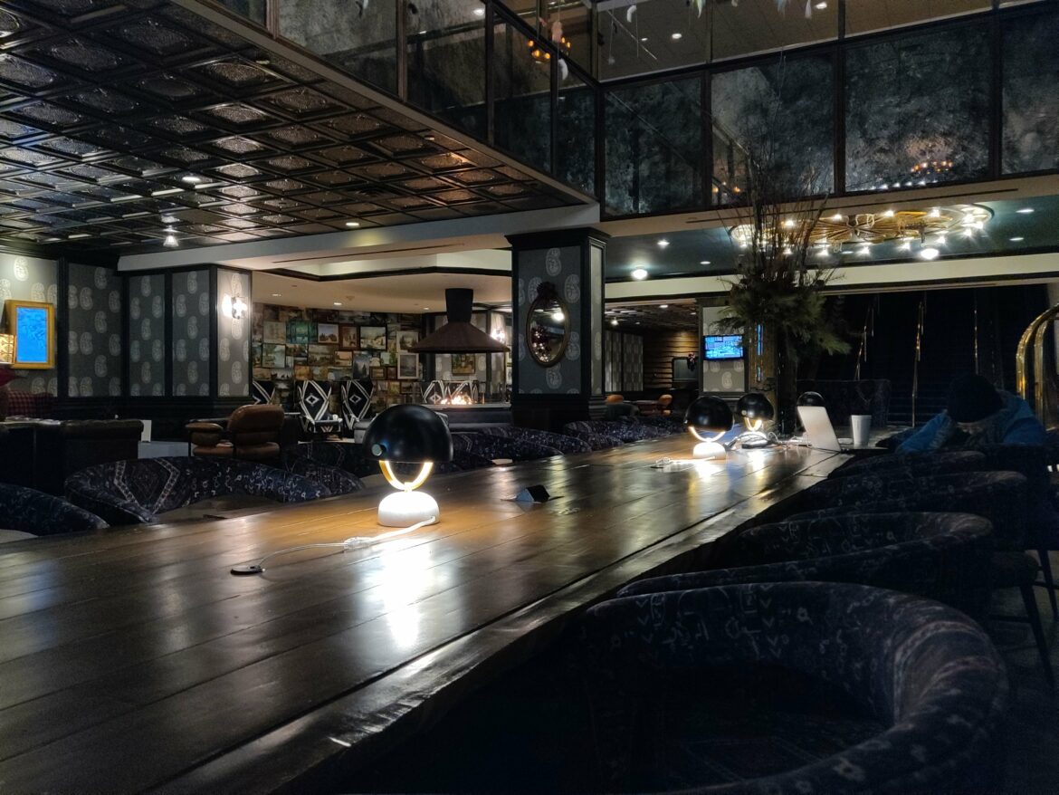 The hilton diamond reserve upgrade shows up in a dimly lit, stylish interior of a lounge or restaurant. There is a long wooden table with modern, spherical lamps placed along its center. The seating around the table consists of patterned chairs. The ceiling has a reflective, textured design, and the walls are decorated with various framed pictures. In the background, there is a cozy seating area with armchairs and a fireplace. A person is seated at the table with a laptop and a drink. The overall ambiance is warm and inviting.