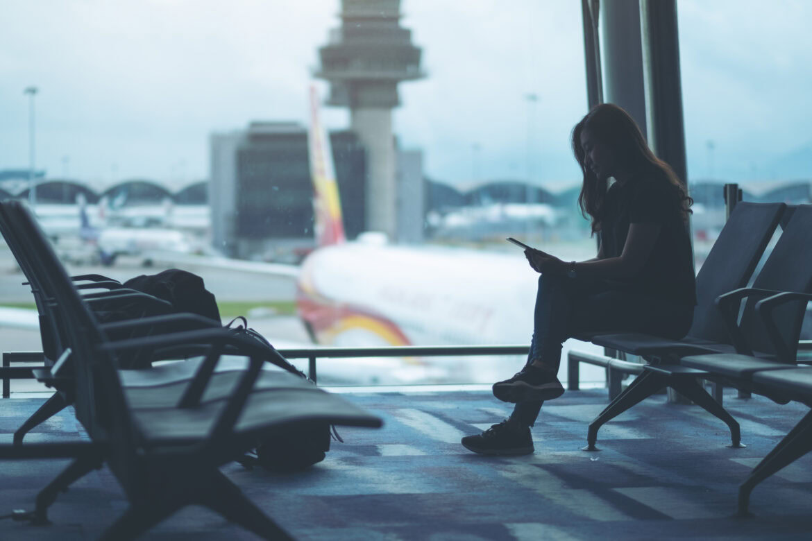 A person is sitting in an airport terminal, looking at a phone. They are silhouetted against a large window through which an airplane and airport structures are visible. The scene conveys a sense of waiting or travel.