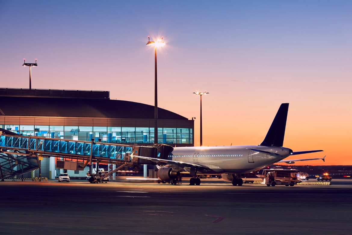 The image shows an airplane parked at an airport gate during sunset. The aircraft is connected to a passenger boarding bridge, and the terminal building is visible in the background with illuminated windows. The sky is a gradient of warm colors, and the tarmac is lit by tall light poles.
