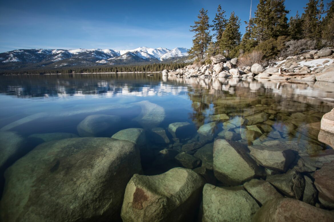 A serene lake with clear water showing rocks beneath the surface. The lake is surrounded by a rocky shoreline and tall pine trees. In the background, there are snow-capped mountains under a clear blue sky.