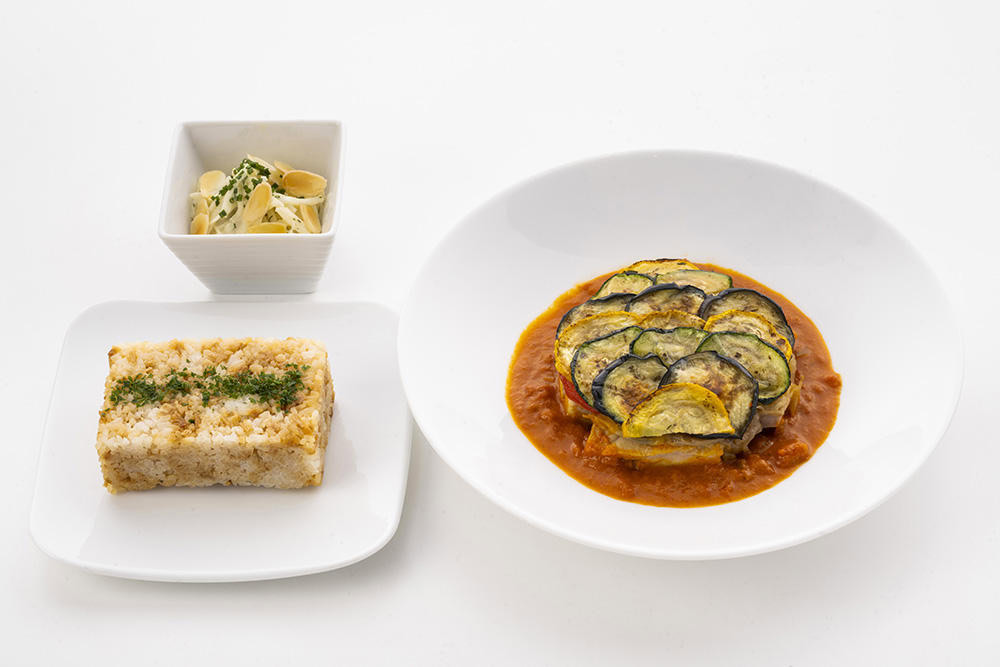 The image shows a neatly arranged meal consisting of three dishes. On the right, there is a white plate with a serving of ratatouille, featuring slices of zucchini and eggplant on a bed of tomato sauce. On the left, there is a square piece of rice with herbs on top, placed on a small white square plate. In the background, there is a small white bowl containing a creamy pasta salad garnished with herbs and almond slices. The dishes are presented on a white surface.