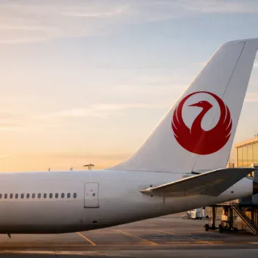 The image shows the tail section of a commercial airplane on the tarmac at an airport during sunset. The tail features a red logo of a stylized bird. In the background, there is a terminal building with large glass windows reflecting the warm light of the setting sun.