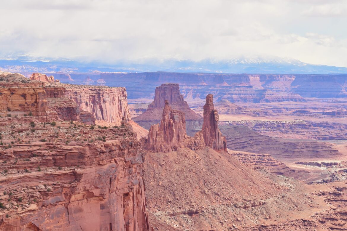 A vast desert landscape featuring towering red rock formations and mesas under a cloudy sky. The foreground shows rugged cliffs with sparse vegetation, while the background reveals layers of canyons and distant mountains. The scene is bathed in soft, natural light, highlighting the textures and colors of the rock formations.