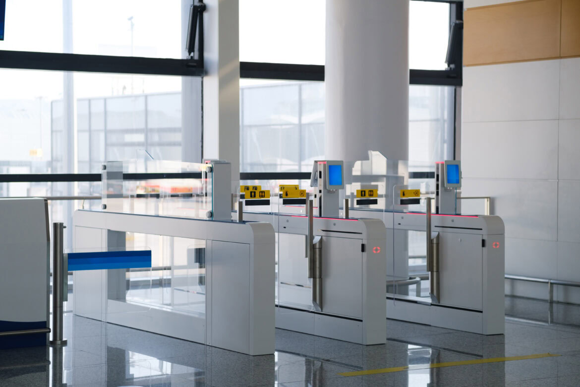 The image shows an airport security checkpoint with automated gates. The gates have glass barriers and are equipped with digital screens and card readers. The area is well-lit with large windows in the background, and the floor is made of polished tiles.