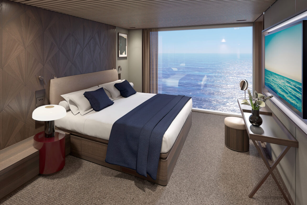 The image shows your Norwegian Luna cruise from a modern, elegantly designed bedroom with a large bed featuring white linens and dark blue pillows and a throw. The room has a textured wooden accent wall and a large window offering a view of the ocean. A small side table with a lamp is next to the bed, and a desk with a vase of flowers and a small mirror is positioned against another wall. The room is well-lit and has a serene, luxurious atmosphere.