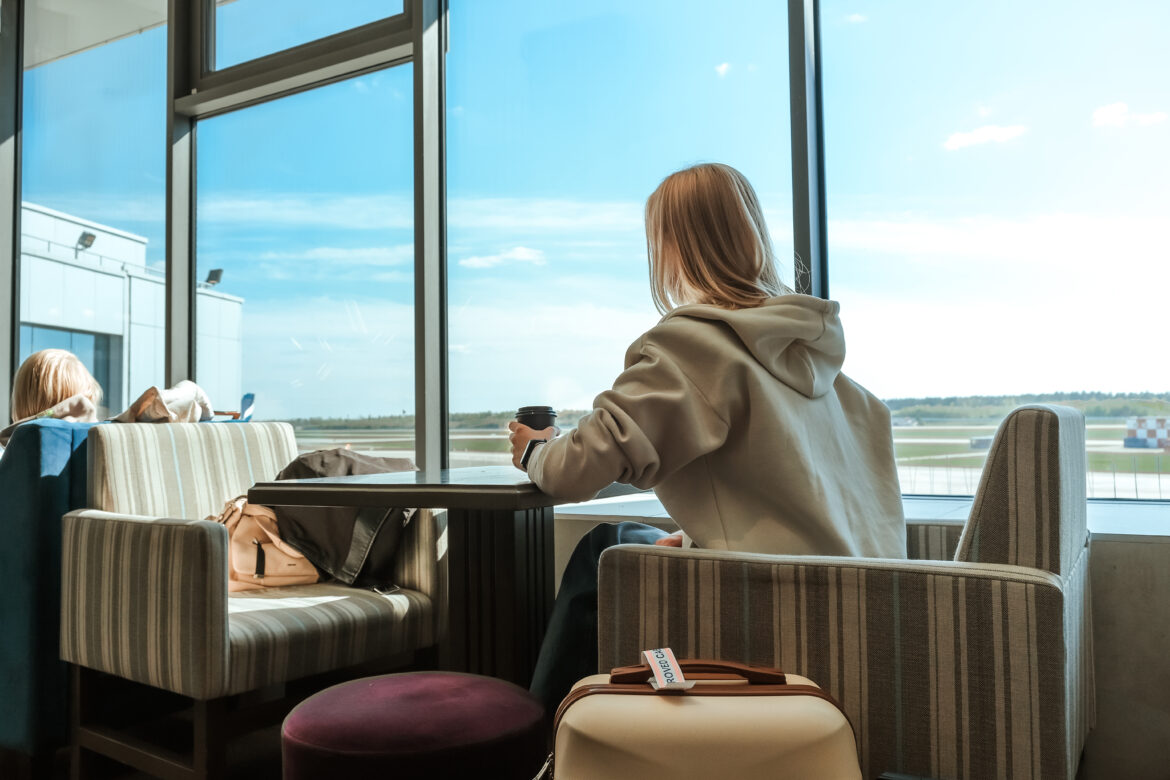 A person with long hair is sitting in an airport lounge, looking out of a large window at the runway. They are wearing a light-colored hoodie and holding a coffee cup. There is a suitcase with a luggage tag on a nearby chair, and another person is sitting in the background. The sky outside is clear and blue.