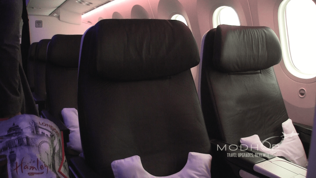 The image shows the interior of an airplane cabin with two adjacent black leather seats. Each seat has a white pillow placed on it. The cabin is dimly lit with a soft pink light, and there are windows visible on the right side. A person is partially visible on the left, holding a bag with a design featuring landmarks and the word "London."